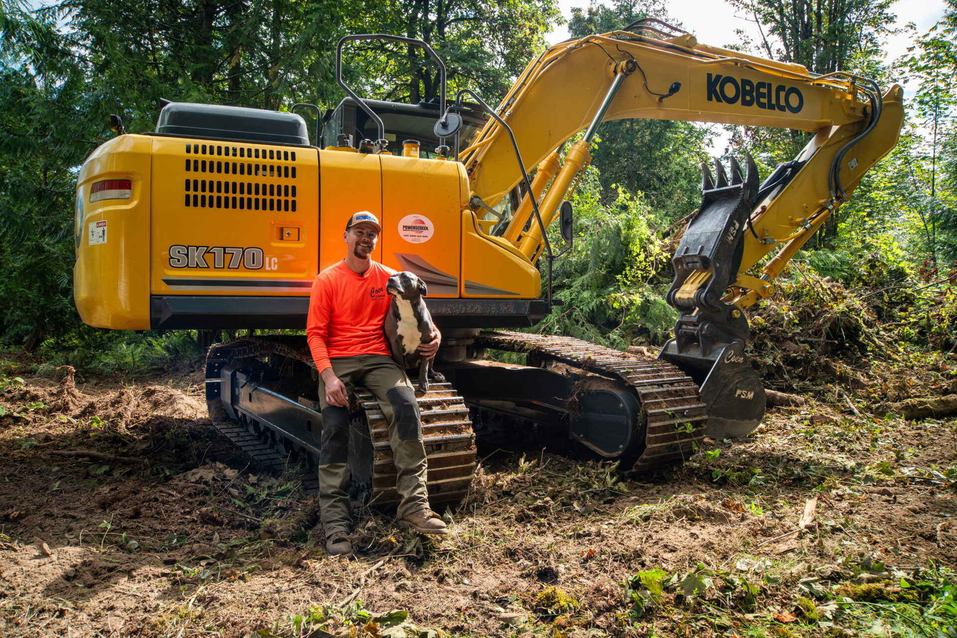 Man in a safety shirt with his dog sitting on his Kobelco SK170LC-11 excavator in the woods
