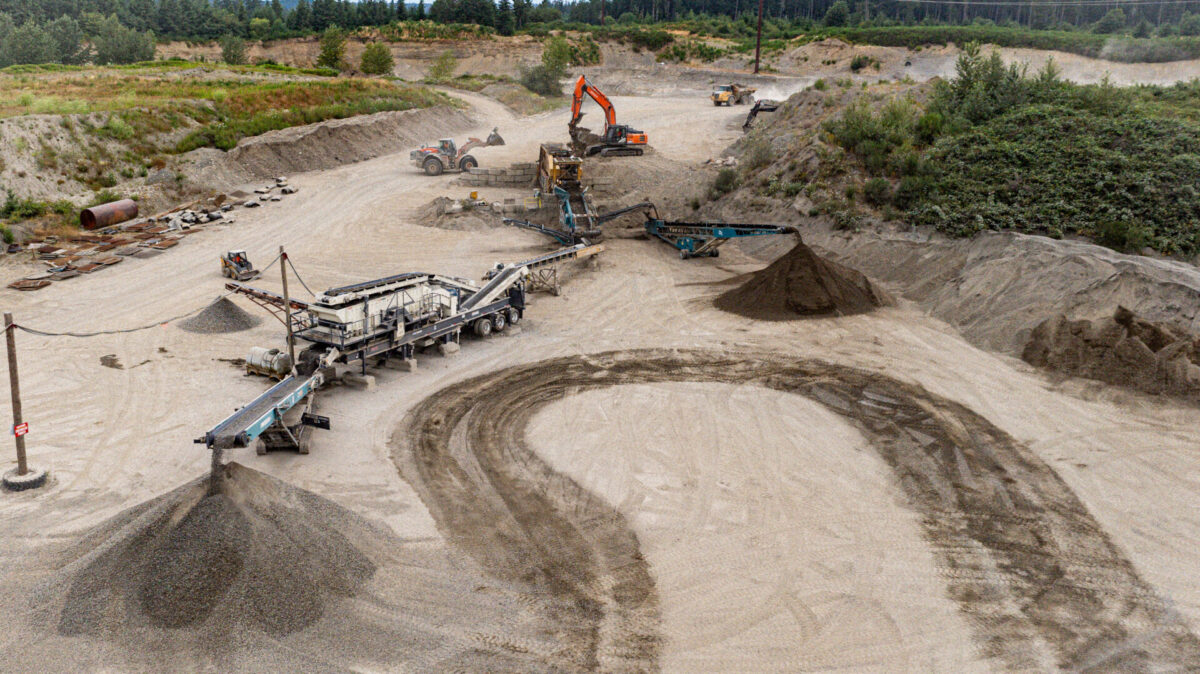 Aerial view of a complete crushing and screening spread processing aggregate at a quarry, featuring a Cedarapids portable cone crusher and conveyor stackers operating on a large sand and gravel site.