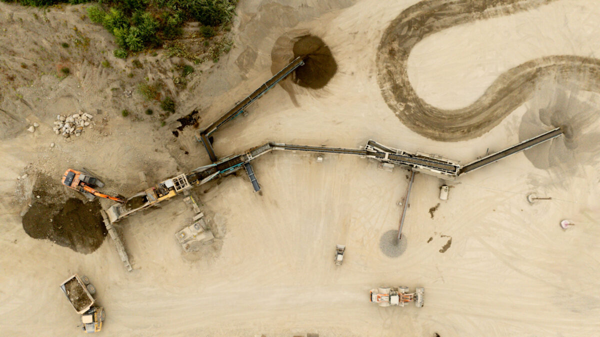 Top-down drone photo of a portable crushing and screening setup at a sand and gravel pit, showing conveyors feeding multiple stockpiles for aggregate production.