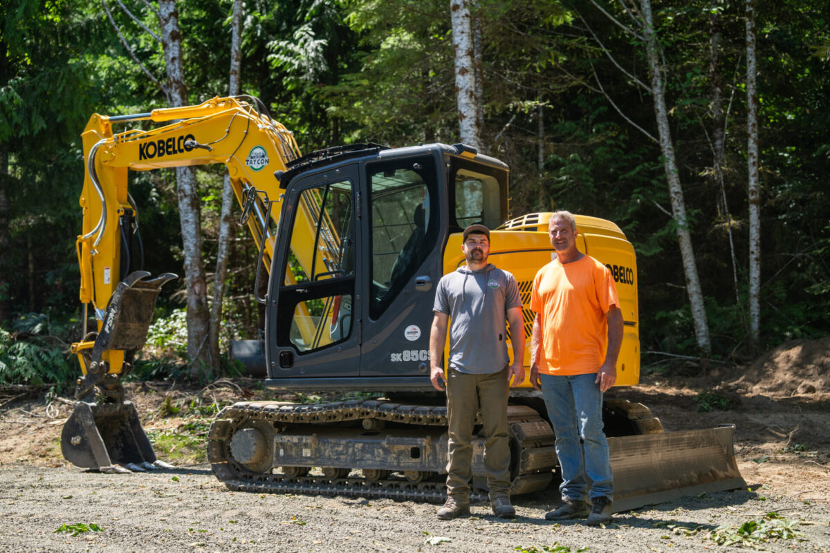 Two men smiling in front of Kobelco SK85 machine